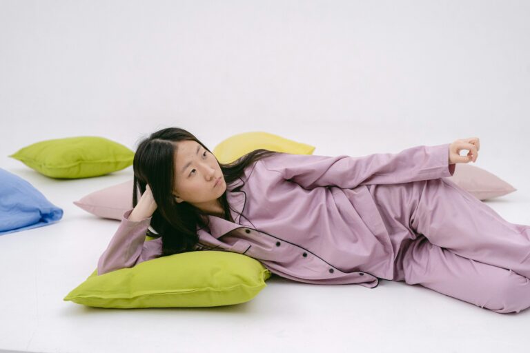 A young woman in pink pajamas lying on colorful pillows, looking thoughtful.